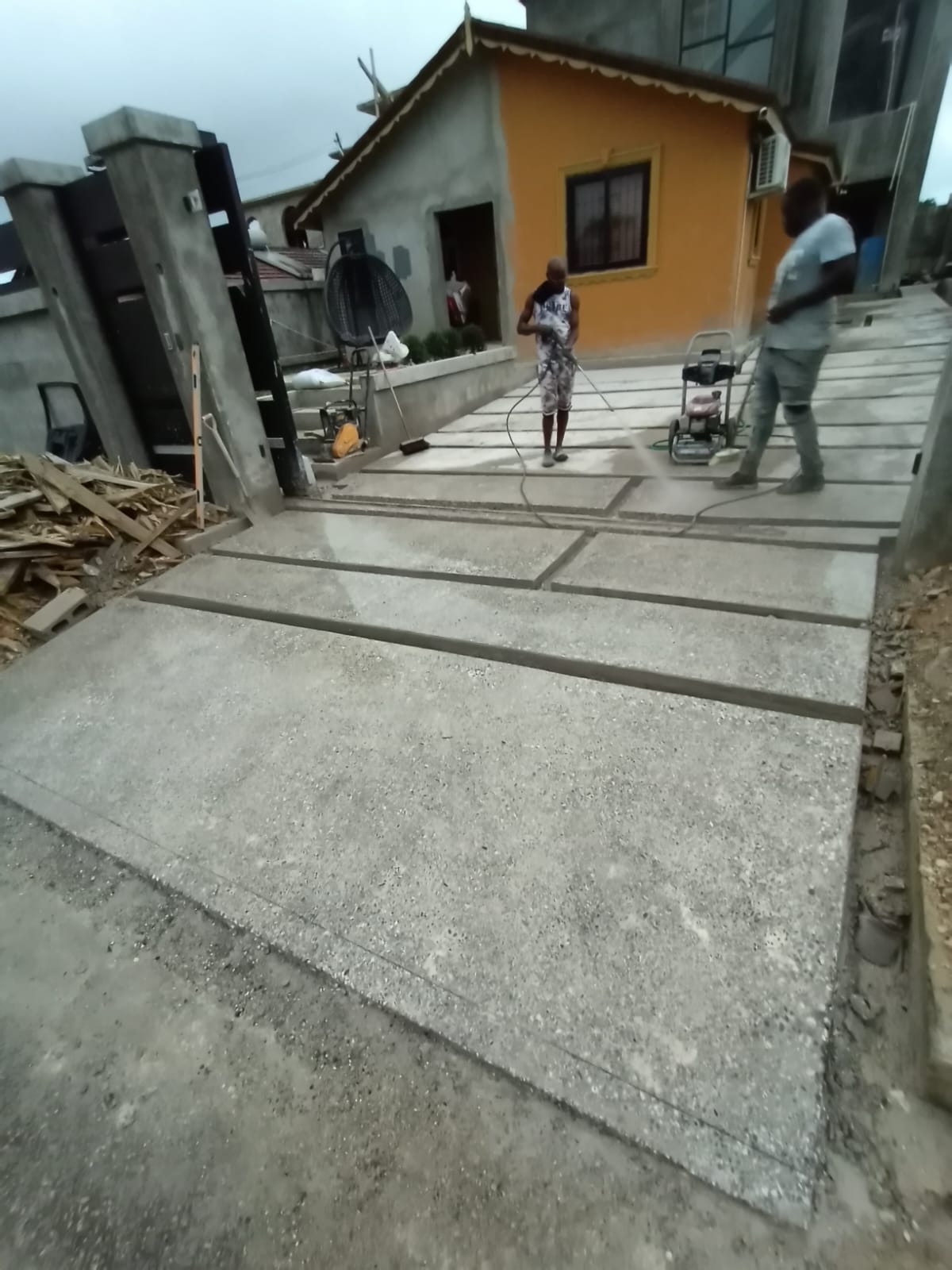 Full house-facing view of the Montego Bay driveway: rectangular honed concrete slabs lead toward the orange house, with the crew finishing the surface.